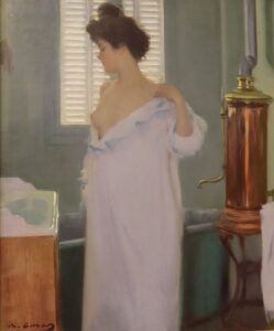 Una dona es troba en una habitació molt il·luminada, parcialment coberta per una túnica, mirant cap a la finestra. Un escalfador daigua de coure és visible al fons. Lescenari suggereix una escena de principis del segle XX.