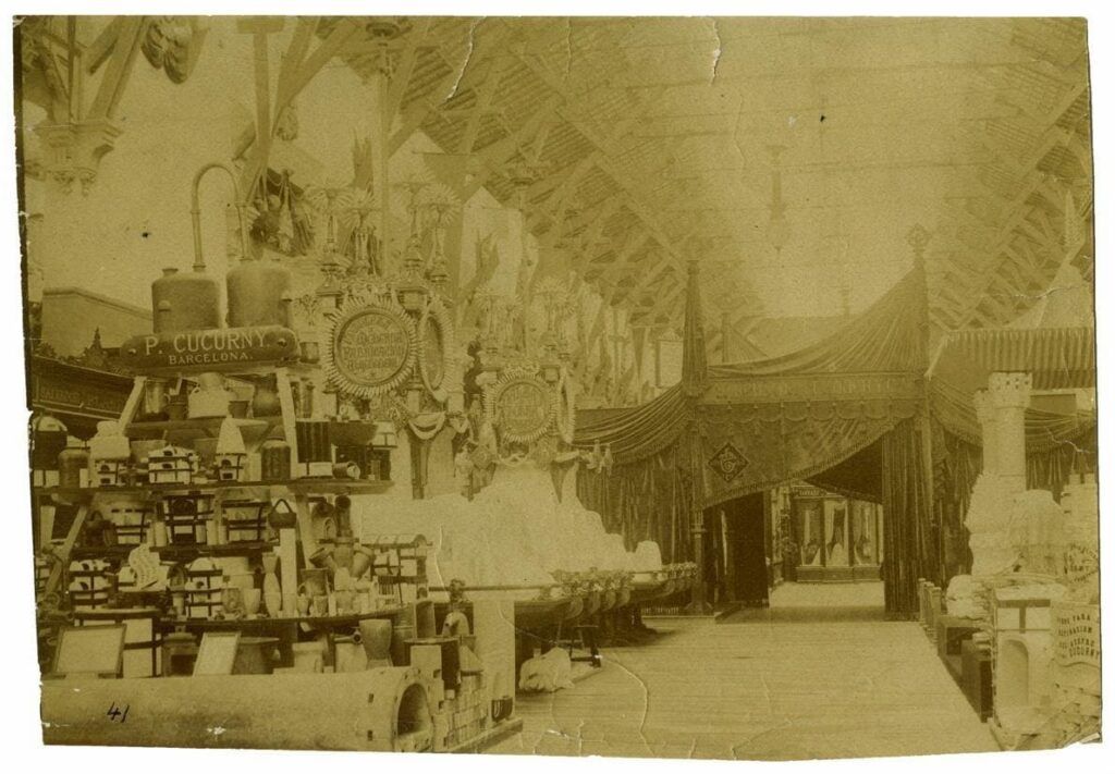 Fotografía en tono sepia de una exposición en interiores con elementos ornamentales, cortinas decorativas y diversos artículos en estanterías. El entorno parece ser de finales del siglo XIX o principios del XX, con detalles arquitectónicos y carteles visibles.