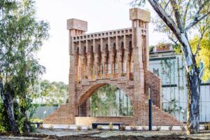 A laire lliure hi ha una gran escultura de maó que sassembla a un pont. Presenta arcs i torres rectangulars, envoltades darbres i vegetació. El cel és clar i brillant, potenciant els tons terrosos de lestructura.