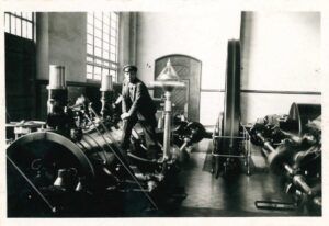 Una foto en blanc i negre dun home amb gorra, dret al costat duna gran maquinària industrial en una fàbrica. Sembla que està operant o inspeccionant lequip, amb tubs i un gran volant visible al seu voltant.