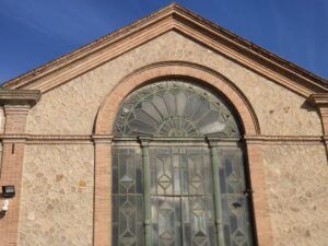 La façana dun edifici històric presenta un gran vitrall darc amb dissenys geomètrics complexos, situat en un mur de pedra. Lestructura té una part superior triangular i la llum del sol projecta ombres suaus a la superfície.