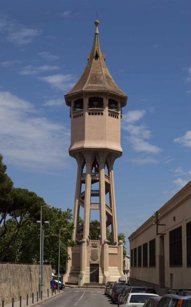 Torre daigua alta i estreta amb teulada apuntada i arcs oberts, envoltada darbres i edificis a banda i banda. Els cotxes estan aparcats al llarg del carrer que hi porta. Cel blau amb núvols dispersos a sobre.