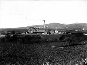 Fotografia en blanc i negre duna gran fàbrica industrial amb chimenees que emeten fum. La fàbrica està envoltada de camps oberts i hi ha muntanyes visibles al fons.