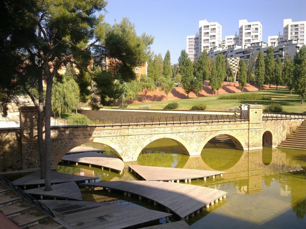 Un pont de pedra sobrepassa un estany tranquil amb camins de fusta a sota. Al fons, hi ha arbres i edificis dapartaments moderns sota un cel blau clar.