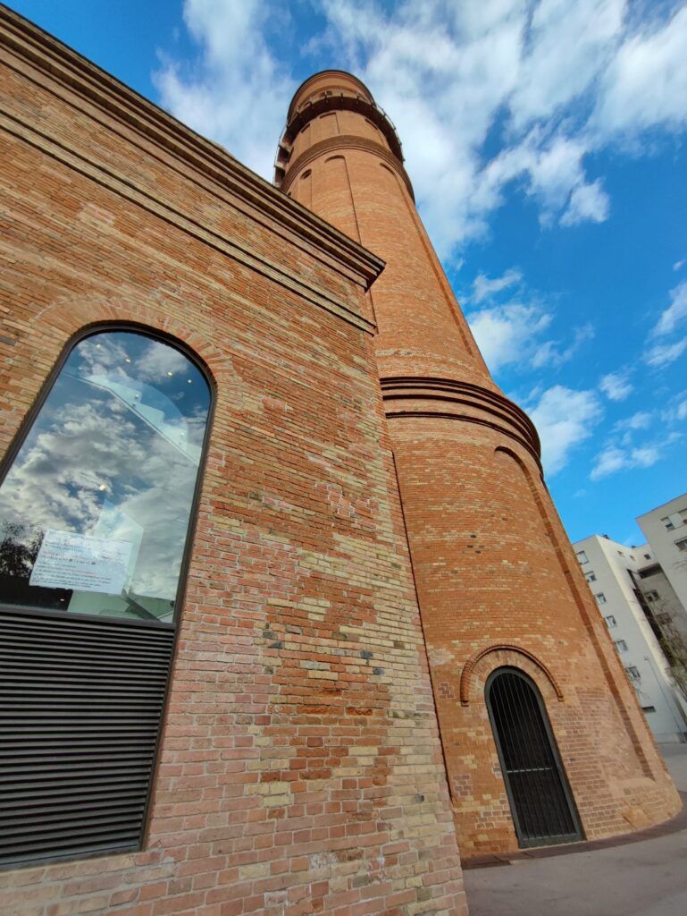 Una torre alta de maó amb finestres darc salça sota un cel parcialment ennuvolat. La seva estructura càlida i taronja contrasta amb el cel blau. A prop, a lesquerra es veu un edifici amb maó semblant.