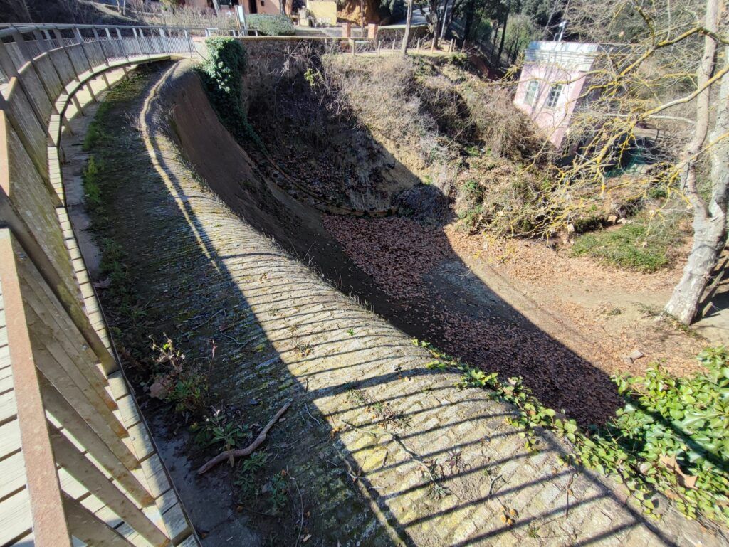 Un aqüeducte de pedra seca i corbada folrada de fulles, que porta cap avall i vorejada per una barana de fusta i arbres. La llum del sol projecta ombres a lestructura. Un petit edifici rosa és visible al fons entre el fullatge.