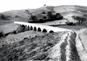 Una foto en blanc i negre dun llarg aqüeducte arquejat que sestén per un paisatge rural i muntanyós. Lestructura presenta múltiples arcs, amb camps i vegetació escassa a banda i banda, sota un cel clar.