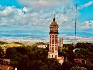 Una torre alta i ornamentada salça enmig duna vegetació exuberant i uns quants edificis més petits, amb un teló de fons de turons ondulants i un cel blau ennuvolat. Una antena alta i esvelta és visible a prop.