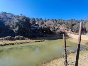 Una escena tranquil·la dun petit estany envoltat darbres i arbustos sota un cel blau clar. En primer pla hi ha una tanca amb pals metàl·lics, mentre que es poden veure cases i arbres en un vessant llunyà.