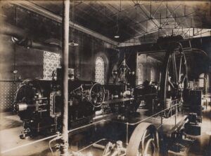 Un interior industrial vintage amb maquinària i equips mecànics grans, amb finestres ornamentades i sostres alts. Lespai està il·luminat per la llum natural que es filtra per les finestres.