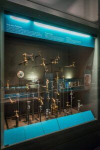 Una exposició del museu titulada Una mar daixetes mostra diverses aixetes metàl·liques disposades de manera creativa sobre canonades. La pantalla sil·lumina amb il·luminació blava i inclou panells de text descriptius a dalt.