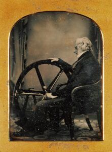Una imatge en tons sèpia dun home gran assegut en una cadira, mirant una gran roda situada davant seu. Sembla pensatiu, amb la mà recolzada al volant. El fons és borrós i la fotografia té un marc vintage.