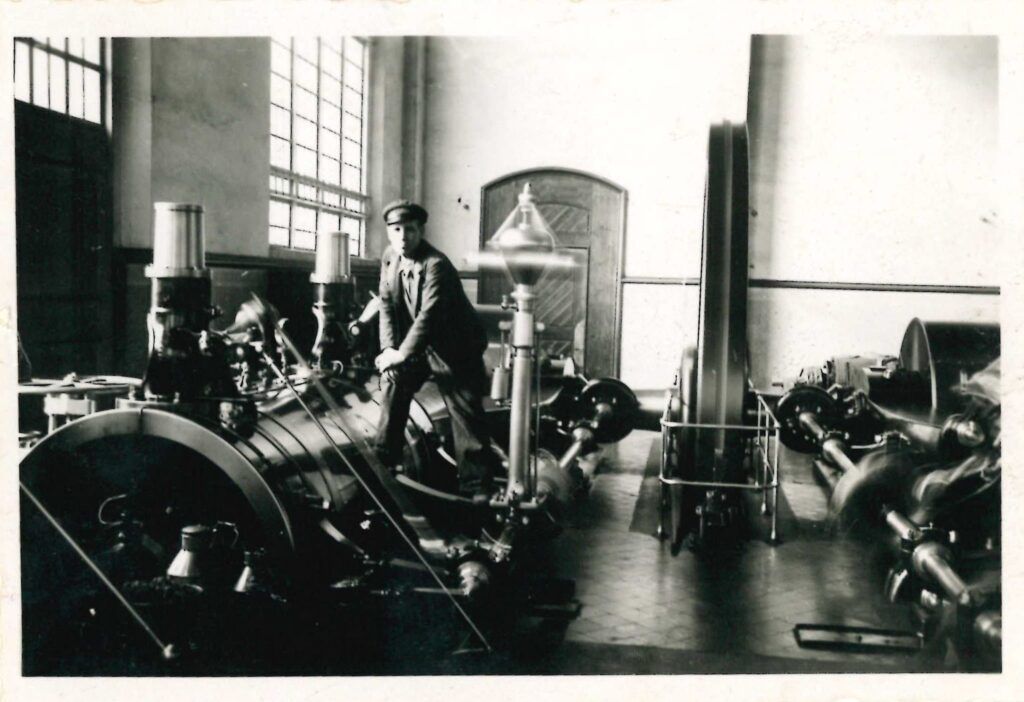 Fotografía en blanco y negro de un hombre con gorra y traje operando maquinaria con grandes ruedas y cilindros dentro de una sala industrial, con grandes ventanas y pisos de baldosas.