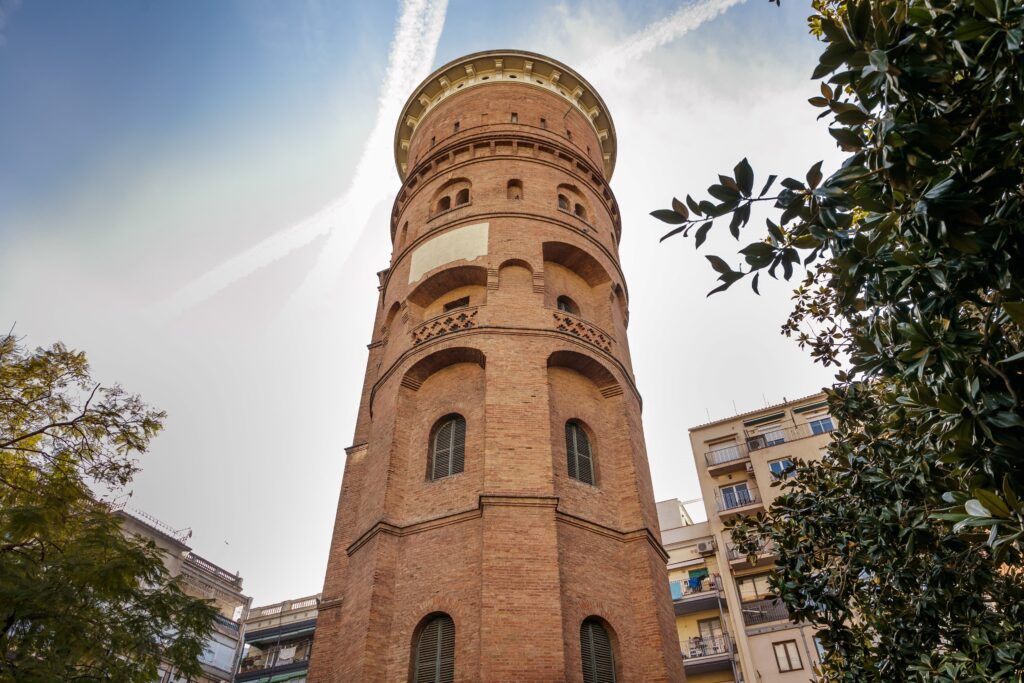 Una torre de ladrillo alta y redonda con ventanas arqueadas se alza contra un cielo azul con nubes tenues. A su alrededor hay árboles y edificios cercanos que capturan una mezcla de naturaleza y arquitectura urbana.