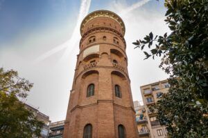 Una torre daigua alta i cilíndrica de maó amb finestres darc i detalls arquitectònics complexos, envoltada darbres frondosos i edificis adjacents sota un cel blau.