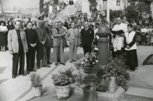 Una foto dèpoca en blanc i negre que mostra un grup dhomes amb vestits formals a prop duna estructura de pedra adornada amb flors. Un sacerdot llegeix un llibre, acompanyat dun jove servidor daltar. Una multitud mira al fons.
