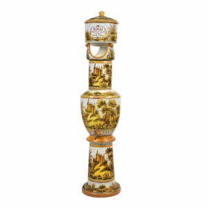 Un dispensador daigua de ceràmica decoratiu amb un disseny paisatgístic vintage, amb arbres i edificis. El dispensador té una estructura alta i estreta amb una espiga prop de la base i una tapa a la part superior amb letiqueta Sinaí.