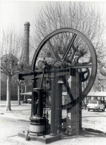 Una gran i antiga màquina de vapor amb un volant d'inèrcia prominent exposat a l'exterior a prop d'arbres sense fulles, una xemeneia de maó, cotxes aparcats i edificis al fons. La imatge és en blanc i negre.