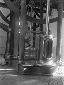 Gran i complexa maquinària de vapor amb columnes verticals, engranatges exposats i components metàl·lics dins d'un edifici industrial amb parets de rajoles i terra enrajolat.