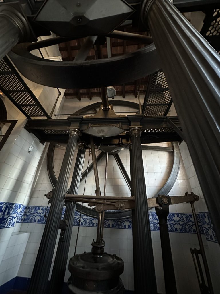 La màquina Alexander amb barres, engranatges i rodes metàl·liques, situada a l'interior amb parets blanques de rajoles amb motius decoratius blaus i un sostre de fusta a sobre.